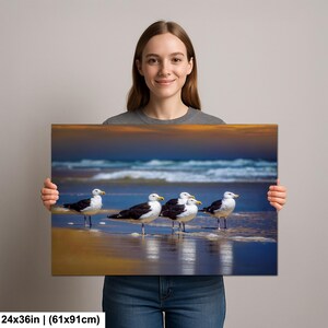Może przedstawiać: Wydruk na pł&oacute;tnie przedstawiający pięć mew stojących na piaszczystej plaży z oceanem w tle. Ptaki mają czarno-białe upierzenie i ż&oacute;łte dzioby. Pł&oacute;tno ma wymiary 61x91cm.