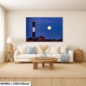 May include: A canvas print of a lighthouse under a full moon. The lighthouse is black and white with a red-roofed building nearby. The print is 40x60 inches (102x152cm) and hangs above a white sofa.