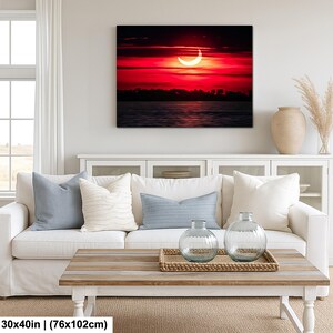 Pode incluir: Impress&atilde;o em tela emoldurada de um vibrante p&ocirc;r do sol vermelho com um sol em forma de crescente sobre a &aacute;gua. A obra de arte, 76x102cm, est&aacute; pendurada acima de um sof&aacute; branco com almofadas azuis. A sala tem uma mesa de centro de madeira.