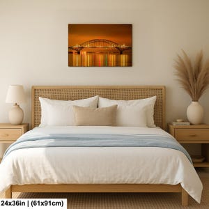 May include: A framed canvas print of a bridge at night, with lights reflecting in the water. The artwork is 24x36 inches. The bridge has an arched design and is illuminated with warm-toned lights against a dark sky.