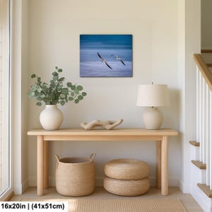 May include: A framed 16x20 inch (41x51cm) print of two seagulls in flight over a blue ocean. The artwork hangs above a light wood console table with a white vase, a lamp, and woven baskets. The room has a neutral color palette.