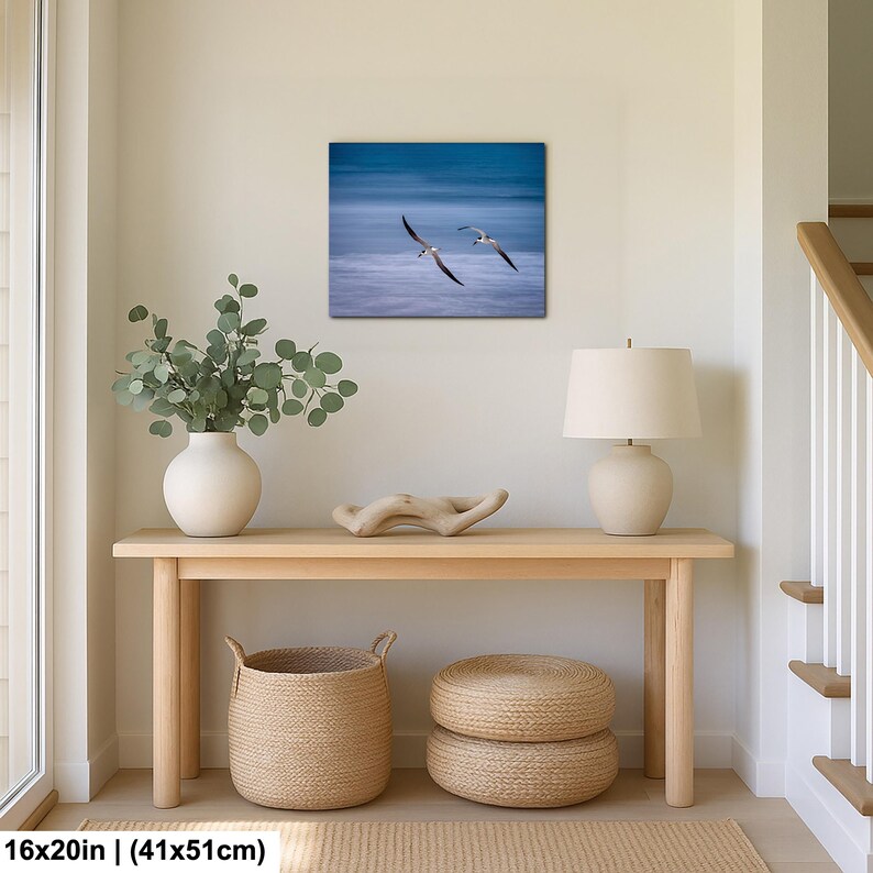 May include: A framed 16x20 inch (41x51cm) print of two seagulls in flight over a blue ocean. The artwork hangs above a light wood console table with a white vase, a lamp, and woven baskets. The room has a neutral color palette.