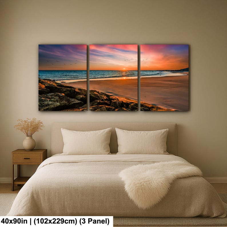 May include: A three-panel canvas print depicting a beach scene at sunset. The sky transitions from pink and purple to orange and yellow, reflecting on the water. The beach has brown sand and dark rocks. The dimensions are 40x90 inches.