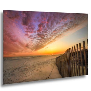 Peut inclure: Paysage de plage avec un coucher de soleil spectaculaire. Le ciel est rempli de nuages roses, oranges et violets. Une cl&ocirc;ture en bois longe le c&ocirc;t&eacute; droit de la plage, avec des empreintes de pas dans le sable.