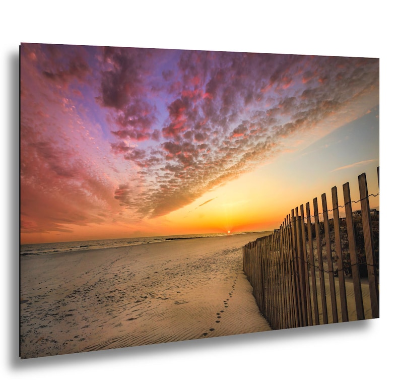 Peut inclure: Paysage de plage avec un coucher de soleil spectaculaire. Le ciel est rempli de nuages roses, oranges et violets. Une cl&ocirc;ture en bois longe le c&ocirc;t&eacute; droit de la plage, avec des empreintes de pas dans le sable.