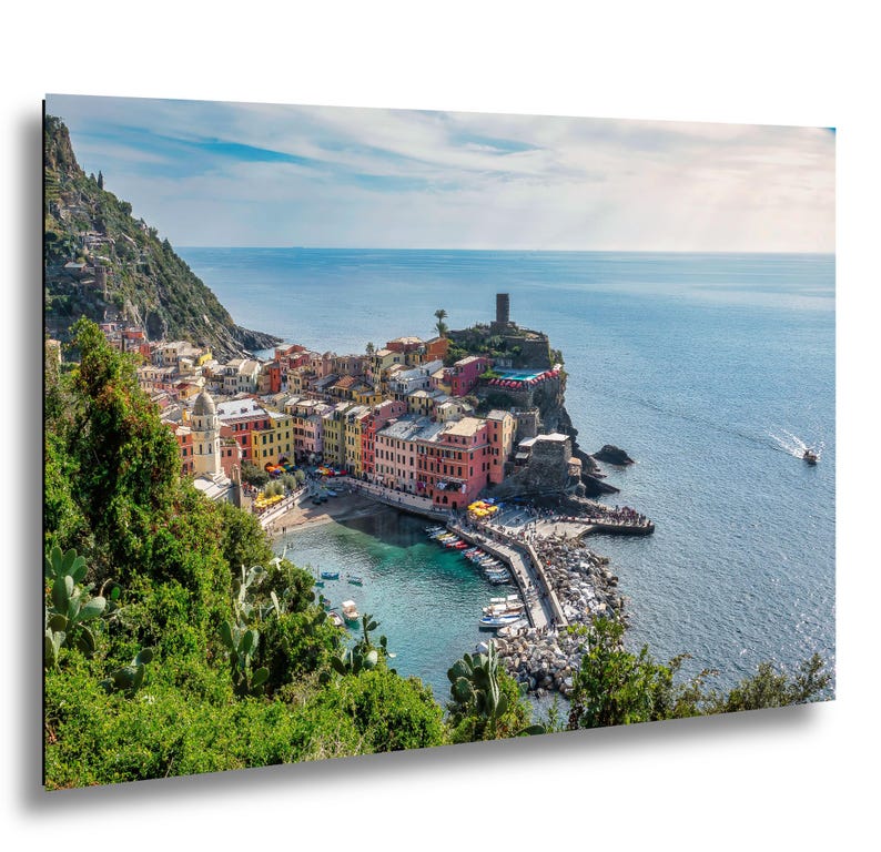 May include: A vibrant coastal town print featuring colorful buildings nestled along a rocky coastline. The image showcases a harbor filled with boats, a stone pier, and a clear blue sea under a bright sky. A picturesque landscape scene.