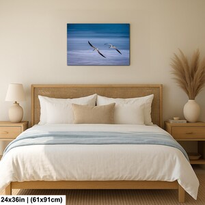 May include: A canvas print featuring two seagulls in flight over a blue ocean. The artwork is rectangular, measuring 24x36 inches. The print is displayed above a bed with a light wood frame and white bedding.