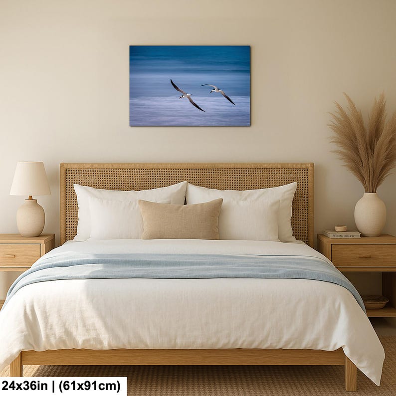 May include: A canvas print featuring two seagulls in flight over a blue ocean. The artwork is rectangular, measuring 24x36 inches. The print is displayed above a bed with a light wood frame and white bedding.