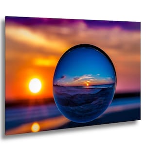 May include: A clear glass sphere positioned in the foreground, refracting an inverted image of a beach sunset. The background features a vibrant orange and purple sky with a bright sun, while the sphere displays a cool blue-toned horizon.