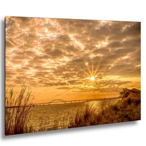 May include: A scenic photograph of a sunset over a body of water, featuring a bridge in the distance. The sky is filled with golden clouds, and the sun is a bright focal point. Tall grasses frame the water's edge.