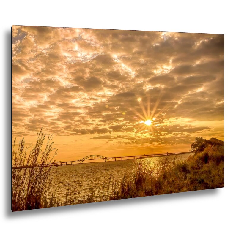 May include: A scenic photograph of a sunset over a body of water, featuring a bridge in the distance. The sky is filled with golden clouds, and the sun is a bright focal point. Tall grasses frame the water's edge.