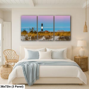 Op de afbeelding: Een drieluik canvas print met een kustlandschap. Het kunstwerk toont een vuurtoren met zwart-witte strepen tegen een roze en blauwe lucht. De afmetingen zijn 91x183 cm.