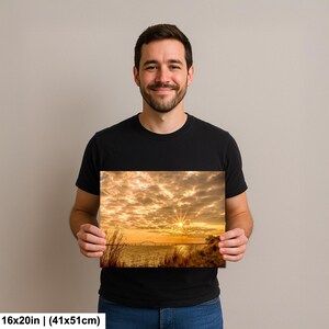 May include: A 16x20 inch (41x51cm) canvas print of a sunset over water, with a bridge in the distance. The sky is filled with golden clouds, and the sun is shining brightly. The print is held by a person.