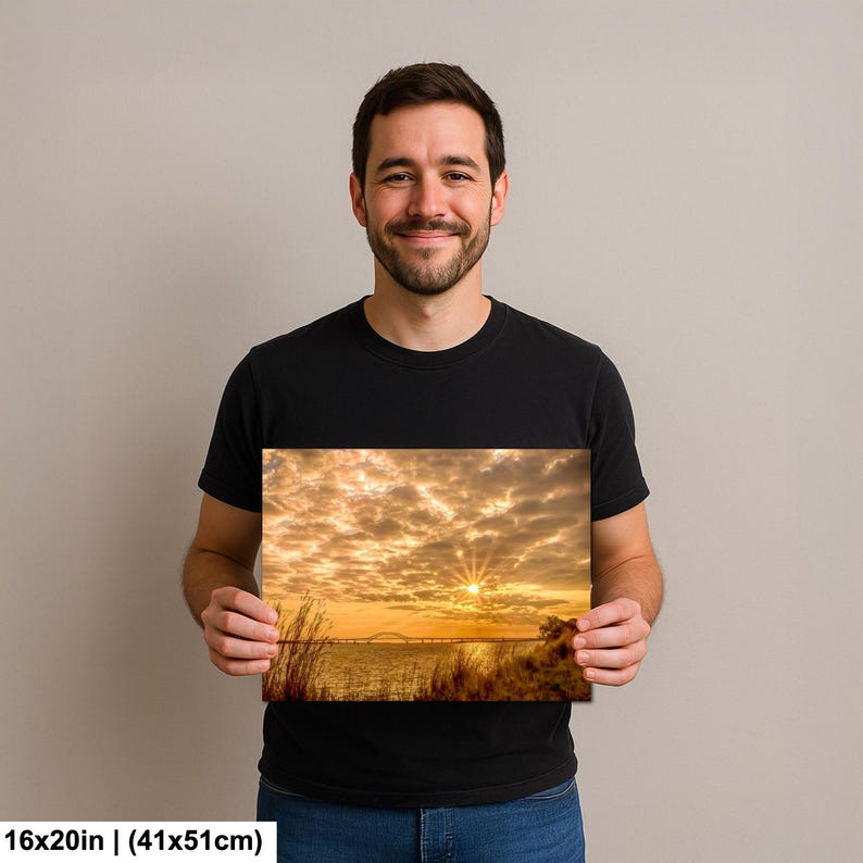 May include: A 16x20 inch (41x51cm) canvas print of a sunset over water, with a bridge in the distance. The sky is filled with golden clouds, and the sun is shining brightly. The print is held by a person.