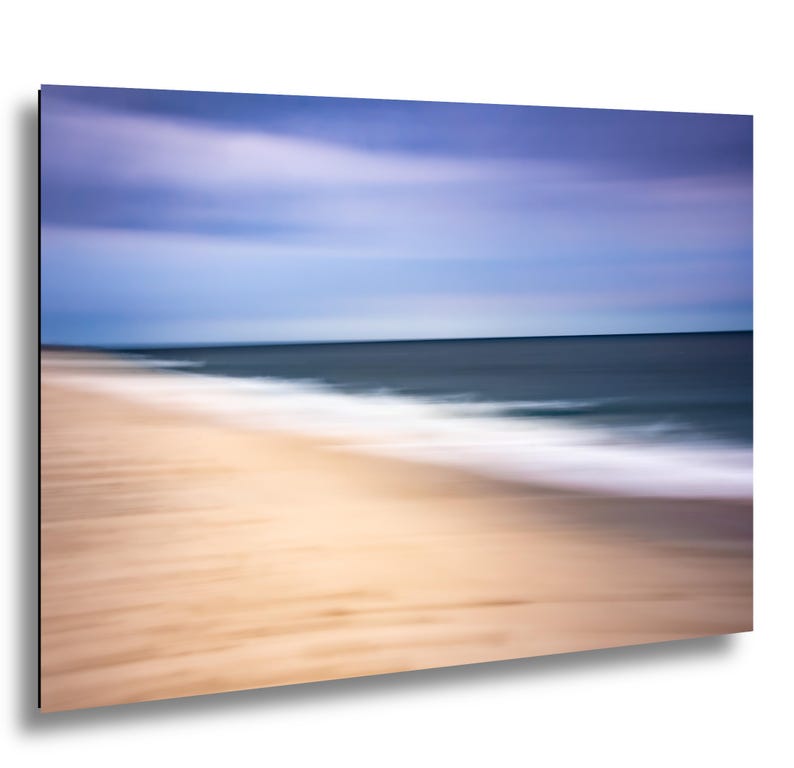 May include: Abstract beach scene with blurred motion. The image features a sandy beach in the foreground, transitioning to the ocean and sky. The colors include shades of tan, blue, and white, creating a sense of tranquility.