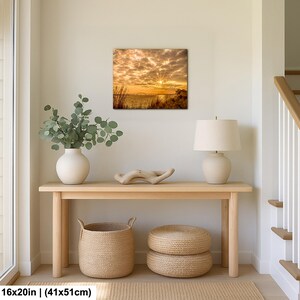 May include: A square canvas print of a sunset over water, measuring 16x20 inches. The image features a vibrant sky with golden and orange hues, and a bright sun. The print is displayed above a wooden console table with decorative items.