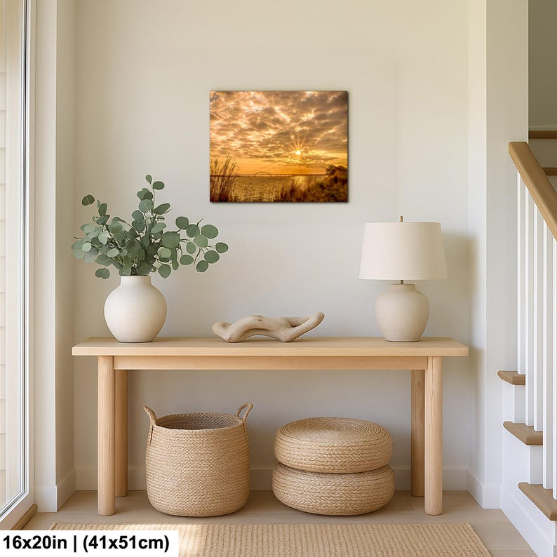 May include: A square canvas print of a sunset over water, measuring 16x20 inches. The image features a vibrant sky with golden and orange hues, and a bright sun. The print is displayed above a wooden console table with decorative items.