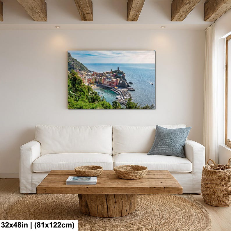 May include: A canvas print of a coastal Italian village, featuring colorful buildings nestled along the shoreline. The artwork, measuring 32x48 inches, captures the vibrant hues of the architecture against the backdrop of the blue sea and sky. The print is displayed above a white sofa.
