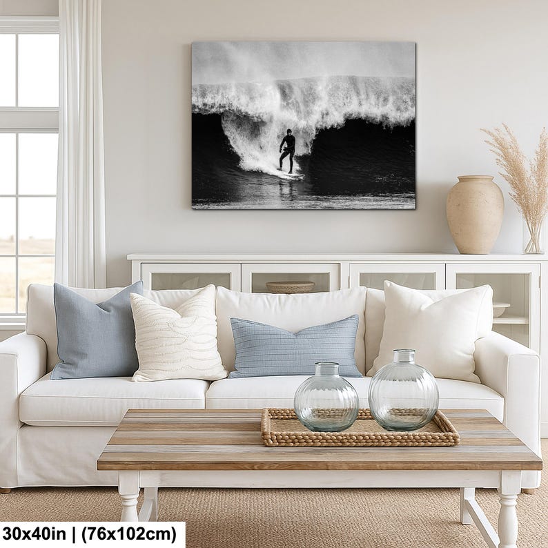 K&ouml;nnte beinhalten: Schwarz-Wei&szlig;-Leinwanddruck eines Surfers auf einer Welle. Das Kunstwerk wird &uuml;ber einem wei&szlig;en Sofa mit blauen und wei&szlig;en Kissen gezeigt. Der Raum verf&uuml;gt au&szlig;erdem &uuml;ber einen Holztisch und dekorative Vasen.