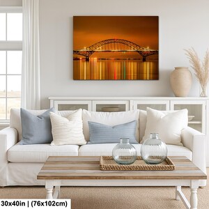 May include: A canvas print of a bridge at night, with lights reflecting in the water. The bridge is arched and has a metal structure. The print is 30x40 inches (76x102cm) and hangs above a white sofa with blue pillows.