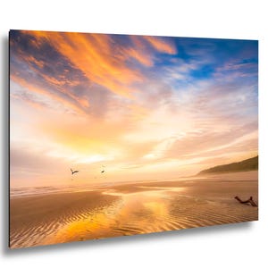 May include: A vibrant beach scene at sunset. The sky is filled with orange, yellow, and blue hues, reflecting on the wet sand. Silhouettes of birds fly across the sky, and a piece of driftwood rests on the shore.