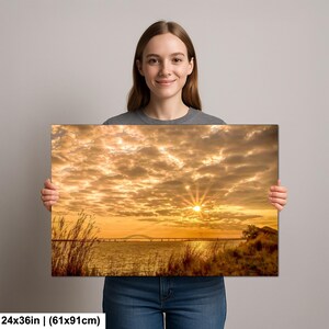 May include: A canvas print featuring a sunset over water with a bridge in the distance. The sky is filled with golden clouds, and the sun is a bright burst of light. The print measures 24x36 inches.