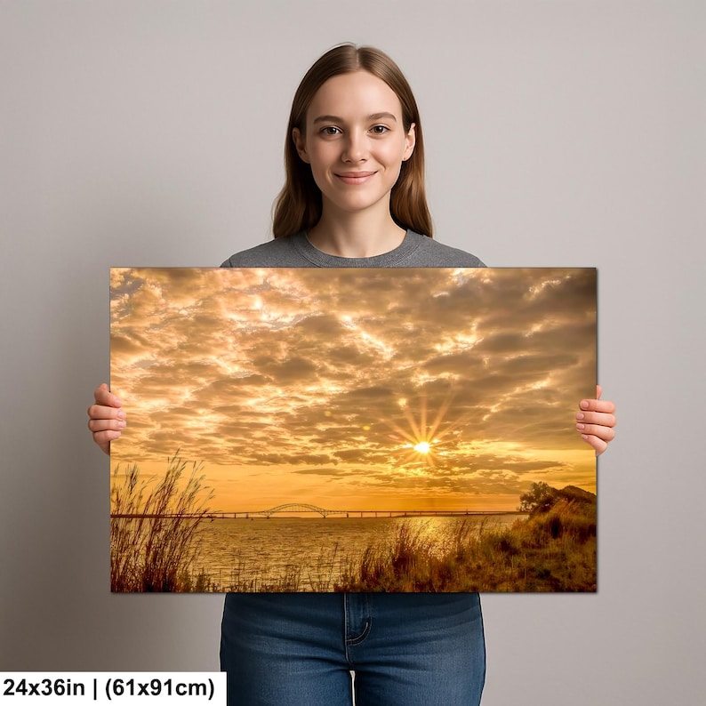 May include: A canvas print featuring a sunset over water with a bridge in the distance. The sky is filled with golden clouds, and the sun is a bright burst of light. The print measures 24x36 inches.