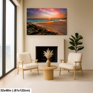 May include: A large canvas print of a beach scene with a vibrant sunset. The artwork, measuring 32x48 inches, features a colorful sky, ocean waves, and a sandy shore. The print hangs above a fireplace, with two armchairs and a coffee table in front.