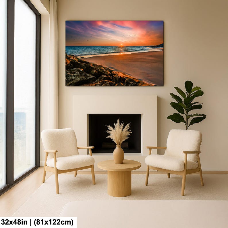 May include: A large canvas print of a beach scene with a vibrant sunset. The artwork, measuring 32x48 inches, features a colorful sky, ocean waves, and a sandy shore. The print hangs above a fireplace, with two armchairs and a coffee table in front.
