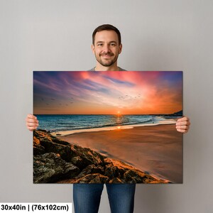 May include: A canvas print depicting a beach scene at sunset. The artwork features a vibrant sky with orange, pink, and purple hues, reflecting on the ocean. The dimensions are 30x40 inches (76x102cm).