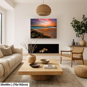 May include: A 30x40 inch (76x102cm) canvas print of a beach scene at sunset. The artwork features a vibrant sky with orange, pink, and purple hues, reflecting on the ocean. The foreground shows rocks and sand.