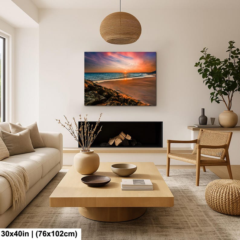 May include: A 30x40 inch (76x102cm) canvas print of a beach scene at sunset. The artwork features a vibrant sky with orange, pink, and purple hues, reflecting on the ocean. The foreground shows rocks and sand.