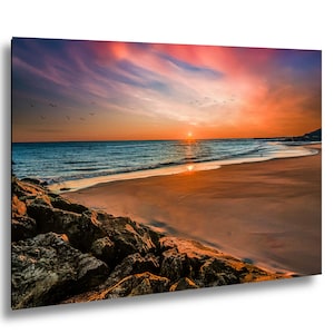 May include: A vibrant print of a beach scene at sunset. The sky is a mix of orange, pink, and blue hues, with a flock of birds flying above the ocean. The foreground features a sandy beach and rocky shoreline.