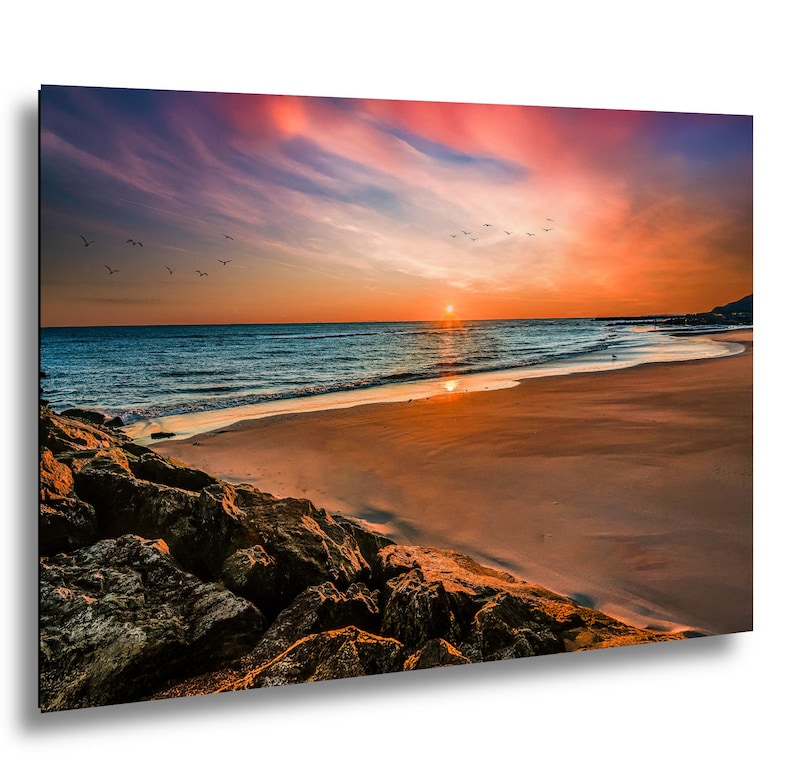 May include: A vibrant print of a beach scene at sunset. The sky is a mix of orange, pink, and blue hues, with a flock of birds flying above the ocean. The foreground features a sandy beach and rocky shoreline.