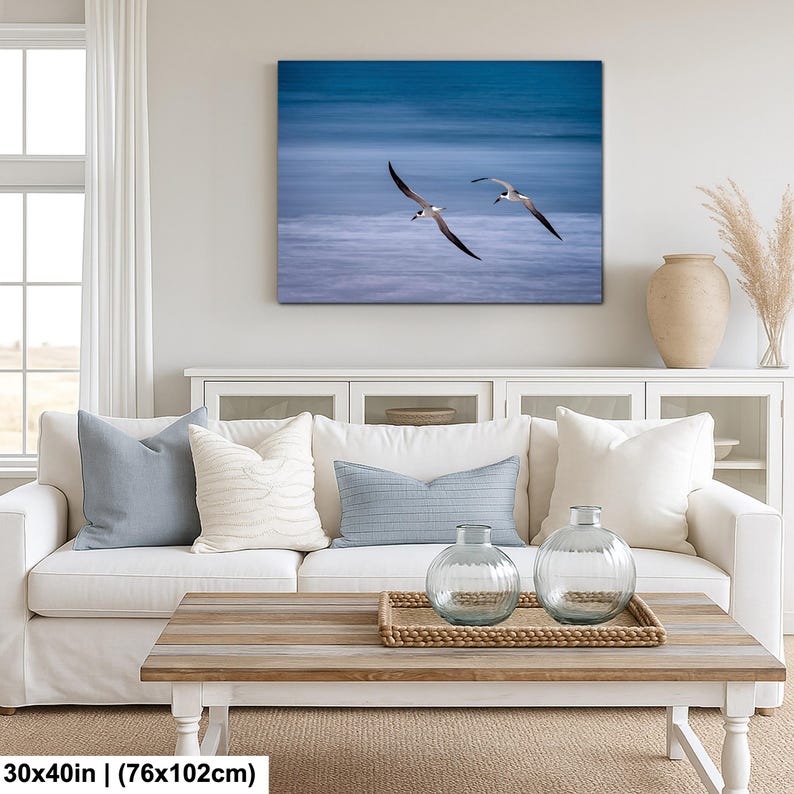 May include: A canvas print featuring two seabirds in flight over a blue ocean. The artwork is displayed above a white sofa with blue and white pillows, a wooden coffee table, and decorative glass vases. The print measures 30x40 inches.