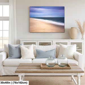 May include: A coastal-themed living room features a white sofa with blue and white pillows, a wooden coffee table, and a large framed canvas art piece depicting a blurred beach scene. Two glass vases sit on a woven tray.