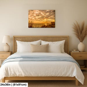 May include: A canvas print of a sunset over water, with a vibrant display of orange and yellow hues in the sky. The artwork, measuring 24x36 inches, is displayed above a bed with white bedding and a light blue throw. The room is decorated in neutral tones.