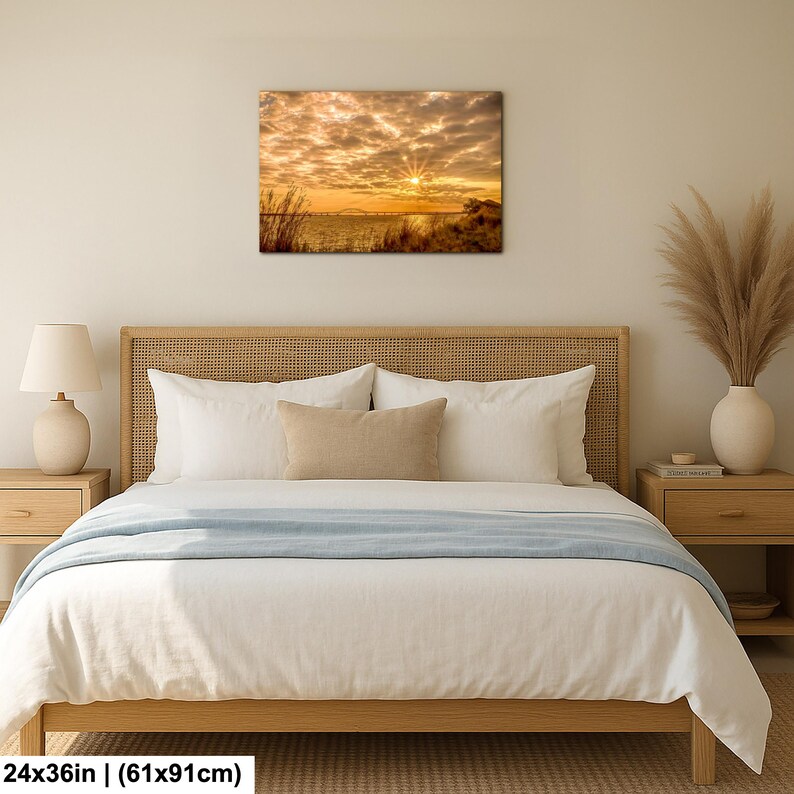 May include: A canvas print of a sunset over water, with a vibrant display of orange and yellow hues in the sky. The artwork, measuring 24x36 inches, is displayed above a bed with white bedding and a light blue throw. The room is decorated in neutral tones.
