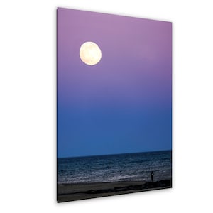 May include: A photograph of a full moon in a gradient sky of purple and blue over a dark ocean. A person stands on the beach. The moon is bright and round, and the ocean has small waves.