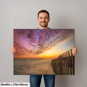 Peut inclure: Impression sur toile d'une sc&egrave;ne de plage au coucher du soleil. Le ciel est rempli de couleurs vives : rose, orange et violet. Une cl&ocirc;ture en bois longe le c&ocirc;t&eacute; droit de la plage. La toile mesure 76x102cm.