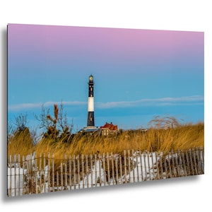 Op de afbeelding: Een schilderachtige print van een vuurtoren met een zwart-witte toren, een gebouw met een rood dak en een houten hek. De lucht gaat over van blauw naar roze, met hoog gras op de voorgrond.