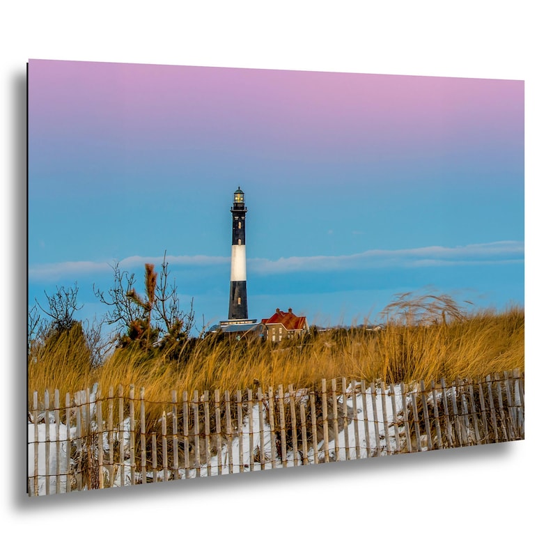 Op de afbeelding: Een schilderachtige print van een vuurtoren met een zwart-witte toren, een gebouw met een rood dak en een houten hek. De lucht gaat over van blauw naar roze, met hoog gras op de voorgrond.