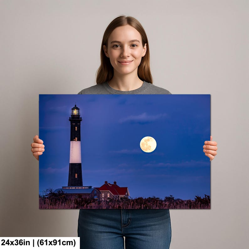 May include: A canvas print featuring a nighttime landscape with a lighthouse and a full moon. The lighthouse is black and white, with a red-roofed building nearby. The moon is bright yellow against a dark blue sky. The print measures 24x36 inches.