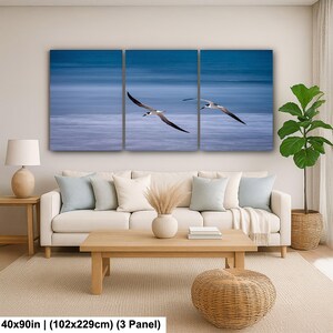 May include: A three-panel canvas print featuring two seabirds in flight against a blurred blue ocean backdrop. The artwork is displayed above a white sofa with light blue pillows, a wooden coffee table, and a woven ottoman. The dimensions are 40x90 inches.
