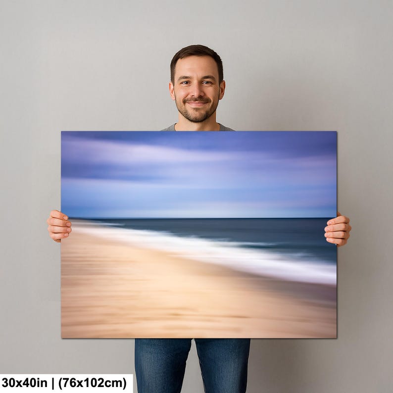 May include: A canvas print featuring a coastal landscape with a blurred effect. The artwork depicts a beach with a sandy shore, ocean waves, and a blue and purple sky. The print measures 30x40 inches.