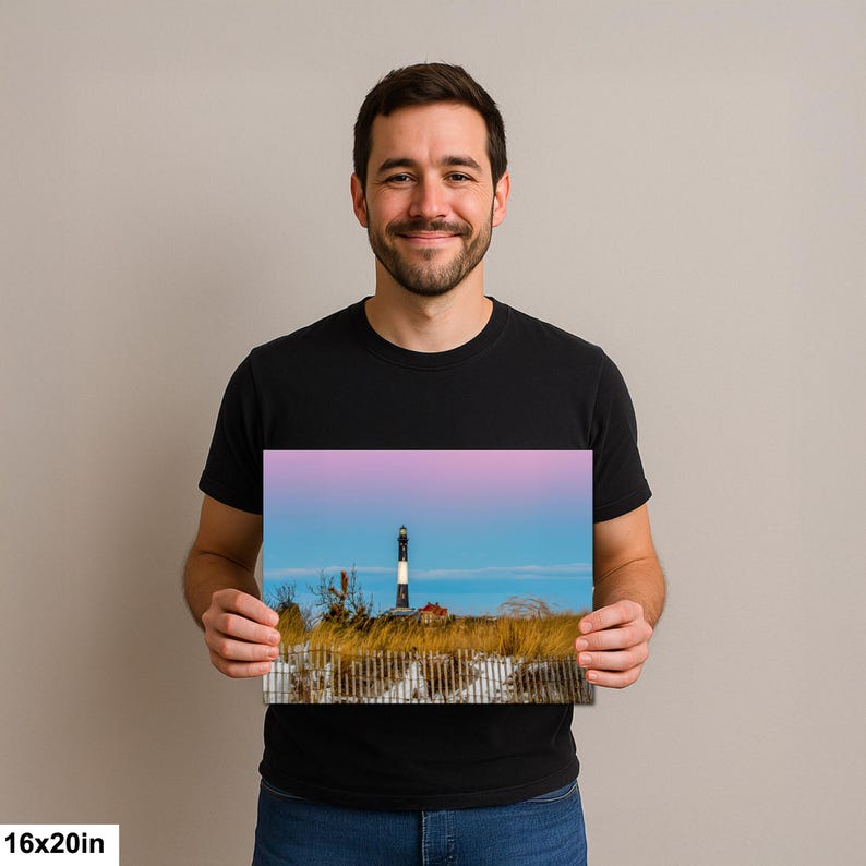 Op de afbeelding: Een 40,6 x 50,8 cm print met een vuurtoren met zwart-witte strepen, tegen een roze en blauwe lucht. De sc&egrave;ne omvat een houten hek, hoog gras en een klein gebouw, waardoor een kustlandschap ontstaat.