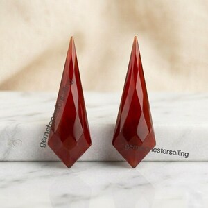 Puede incluir: Dos piedras preciosas de cornalina de color rojo intenso con facetas, en forma de diamantes alargados y puntiagudos. Cada piedra presenta una superficie geométrica multifacetada que refleja la luz, con el texto gemstonesforsalling impreso.
