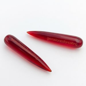 May include: Two elongated, pointed, translucent red glass or gemstone pieces with a smooth, rounded end. Each piece features the text gemstonesforsalling etched in a thin, light-colored font along the side of the object's surface.