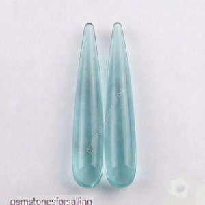 Peut inclure: Deux pierres précieuses bleu clair translucides, en forme de goutte allongée, avec des surfaces lisses et polies. Les pierres sont placées verticalement côte à côte, avec une pointe effilée en haut et une base arrondie en bas. Le texte indique gemstonesforsalling.