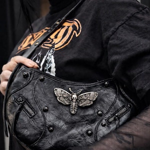 May include: Black leather shoulder bag with a silver moth and skull emblem. The bag has a curved shape, studded details, and a zipper closure. The moth emblem is the focal point of the bag.